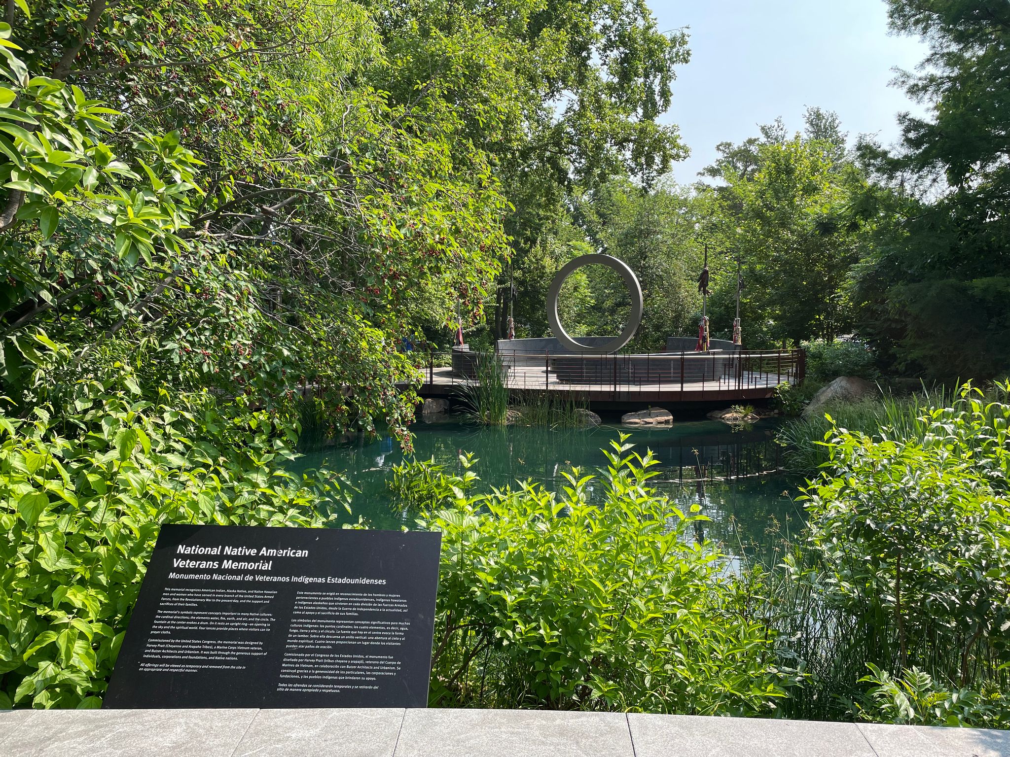 An image of the Nationla Native American Veterans Memorial with green foliage and a pond in the foreground. The memorial, a circular shape, can be seen in the background.