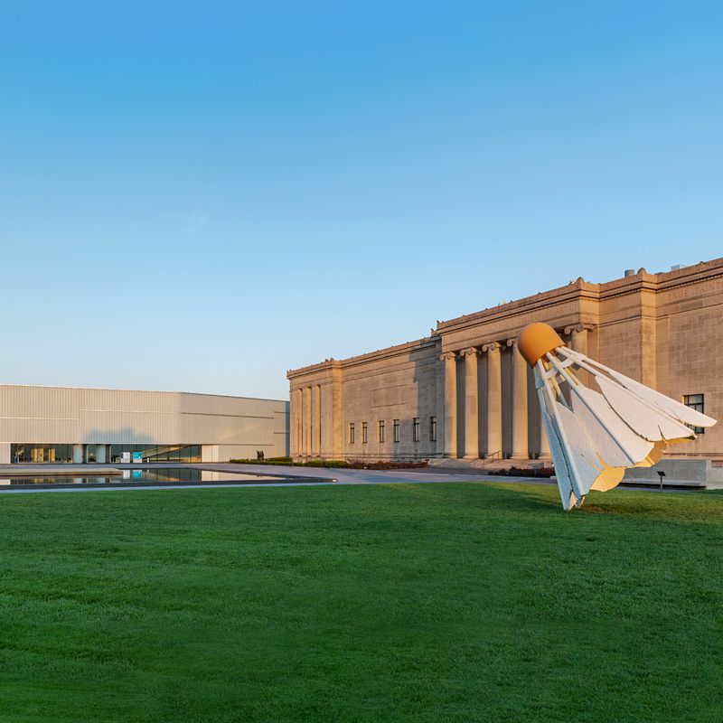 The Nelson-Atkins Museum of Art in Kansas City, featuring its neoclassical façade with large columns and the iconic Shuttlecock sculpture on the lawn in the foreground. The museum's modern Bloch Building extension is visible on the left.