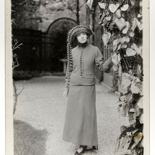 Modèle Chasse : modèle de face en manteau à col montant, boutonnage central et patte et chapeau à longues plumes tombantes, maison Paul Poiret
